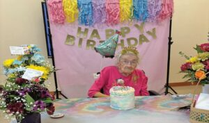 An older woman sitting at a table with a cake in front of her and ballons, streamers, and a banner that says Happy Birthday behind her