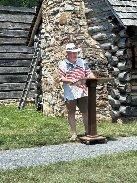 A man speaking at a podium wearing an American flag shirt on a gravel path with a wooden fort behind him