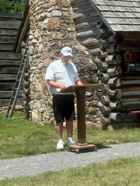 A man speaking at a podium on a gravel path with a wooden fort behind him