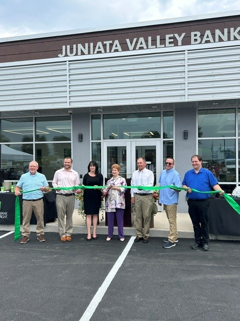 Several people in front of the entrance to a bank holding scissors and a ribbon while posing for a ceremonial ribbon cutting.