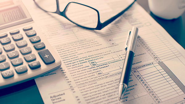 A calculator, glasses, a pen and tax paperwork sitting on a table.]